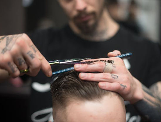 A close-up of a tattooed barber using scissors to trim the hair on a client's head in a stylish barbershop setting.