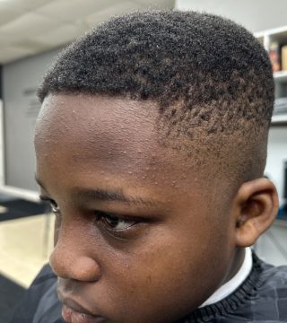 A young boy with a shaved head sits in a barber shop, looking at his reflection in the mirror.
