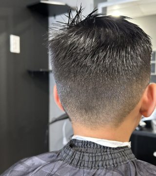 A young man receives a haircut in a modern salon, seated in a stylist's chair with a cape draped over him.