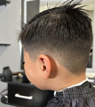 A young boy with a mohawk haircut sits in a barber shop, looking at his reflection in the mirror.