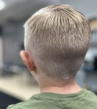 A young boy with a short haircut sits in a salon chair, looking at his reflection in the mirror.