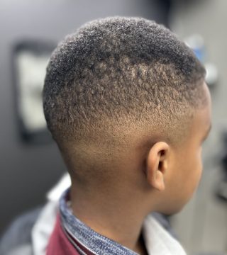 A young boy with a shaved head sits in a barber shop, looking at himself in the mirror with a curious expression.