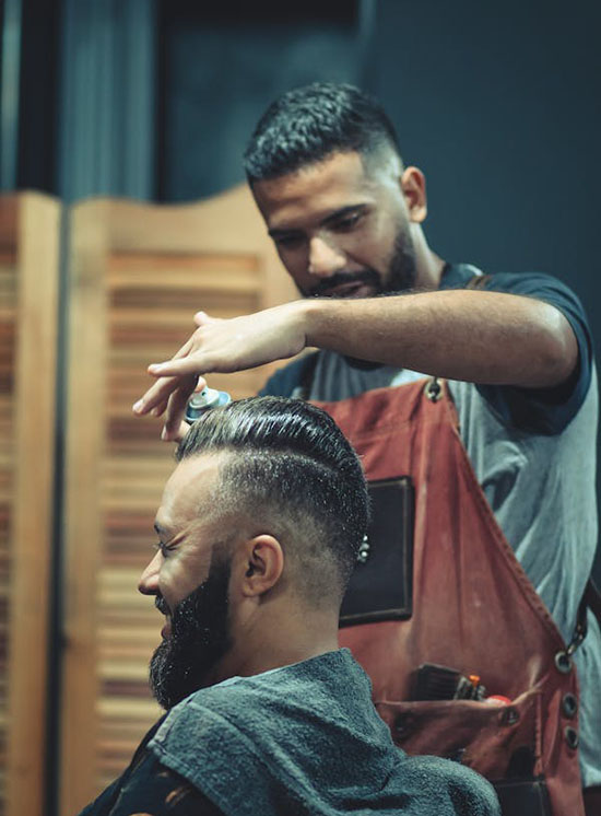 A barber styles a client's hair in a modern salon, surrounded by wooden partitions and equipped with tools for grooming.